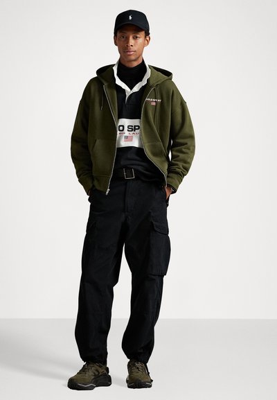 Olive green zip-up hoodie, black cargo pants, and dark sneakers, paired with a black cap and a striped polo shirt with logos.