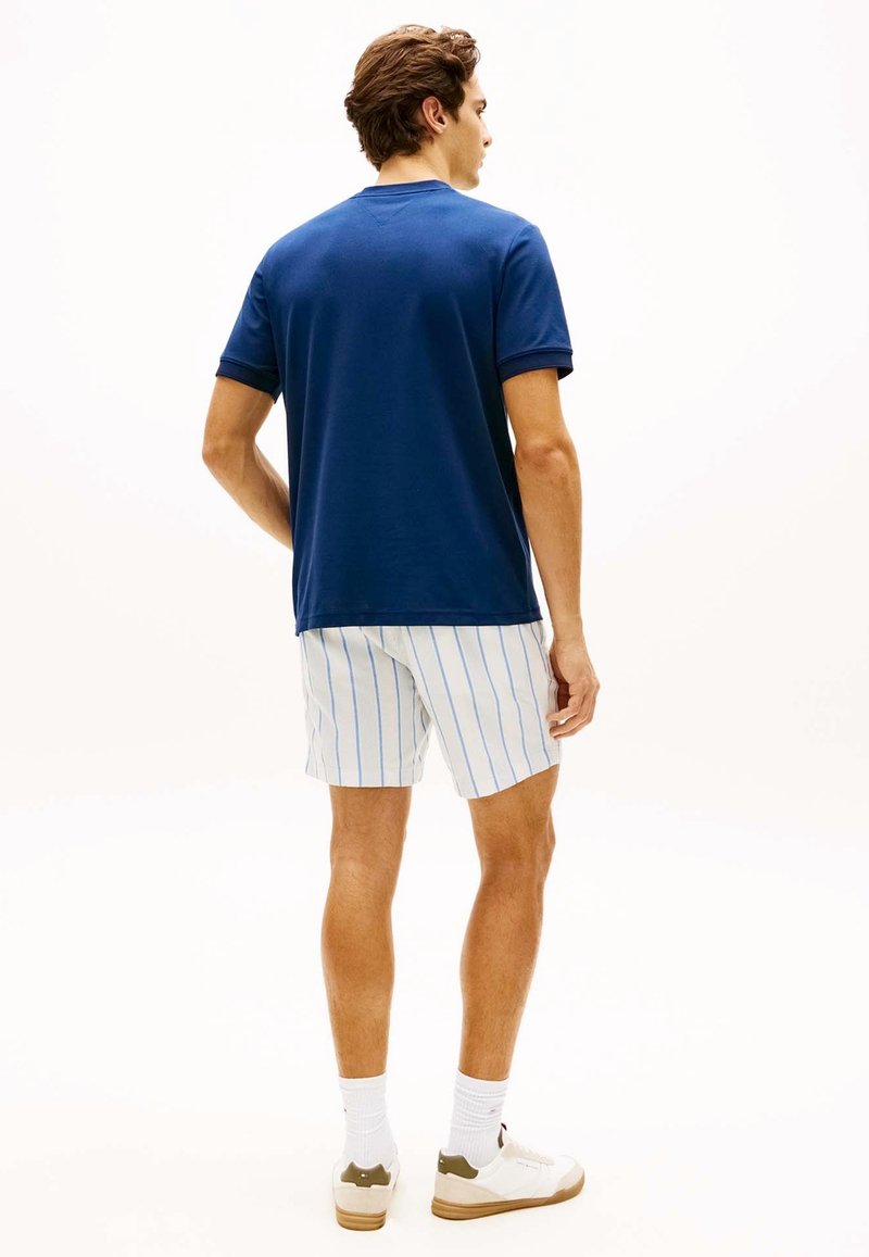 Homme debout de dos portant un t-shirt bleu marine, un short blanc à rayures verticales bleues, des chaussettes blanches et des baskets blanches.