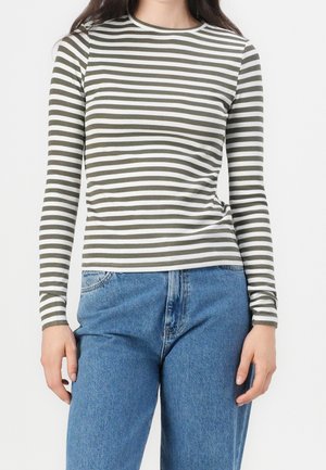 Vrouw draagt een nauwsluitend langarmig shirt met olijfgroene en witte horizontale strepen, gecombineerd met losse blauwe spijkerbroeken, staand tegen een effen achtergrond.