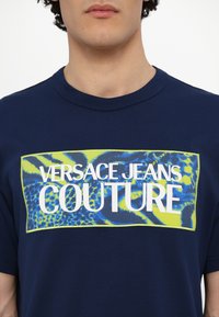 Navy blue cotton t-shirt featuring a colorful graphic panel with "VERSACE JEANS COUTURE" in white text and vibrant patterns in blue and green.