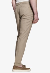 Hombre con pantalones ajustados beige con bolsillos de ojal traseros, combinados con puños remangados y zapatos marrones sin cordones con suelas blancas.