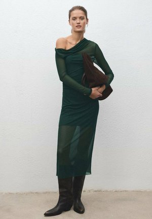 Mujer vestida con un vestido transparente de un solo hombro en verde oscuro, botas negras hasta la rodilla, sosteniendo un gran bolso marrón contra una pared blanca.