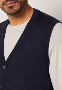 Marinblå ärmlös cardigan med v-ringning, mjuk textur och knappstängning, bärs över en ljus långärmad skjorta.