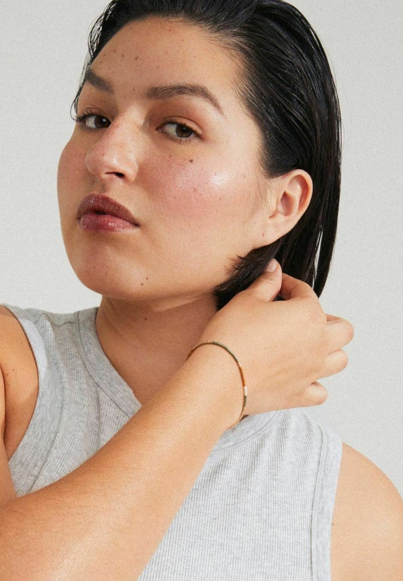 Bracciale color oro con perline verdi e un accento bianco liscio. Indossato su una mano con una maglietta senza maniche grigia, che mette in risalto una finitura testurizzata.