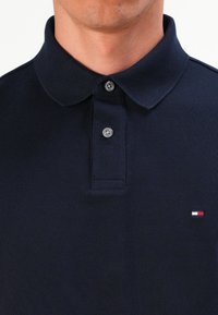 Vyras vilkintis tamsiai mėlyną polo marškinėlį su dviem užsegtais pilkais mygtukais ir mažą raudonai, baltai bei mėlynai spalvotą logotipą ant krūtinės.