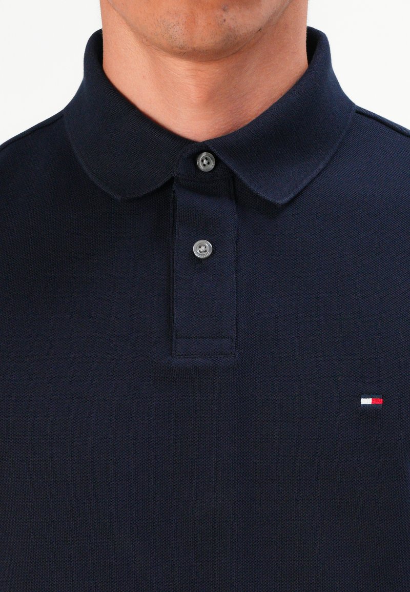 Man med en marinblå pikétröja med två grå knappar fastknäppta och en liten röd, vit och blå logotyp på bröstet.