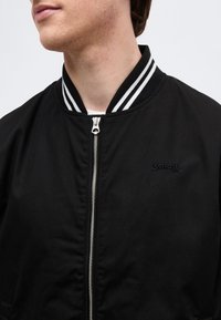 Jeune personne portant une veste noire à fermeture éclair avec un col rayé et un logo discret brodé Schott NYC sur la poitrine.