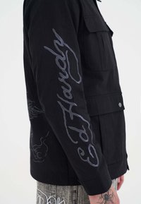 Czarny rękaw kurtki z haftem "Ed Hardy" w ciemnoszarym skrypcie oraz z konturem ręki, noszony z wytatuowanym nadgarstkiem i wzorzystymi spodniami.