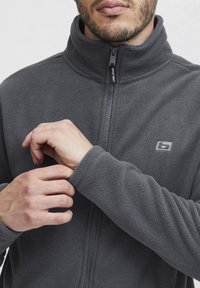 Gray fleece jacket with a high collar and front zipper. Features a logo on the left chest, soft texture, and fitted cuffs.