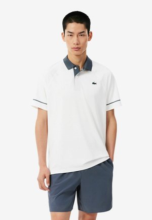 Mann trägt weißes kurzärmeliges Polo mit dunklem Kragen und grauen Shorts, steht vor einfarbigem Hintergrund.