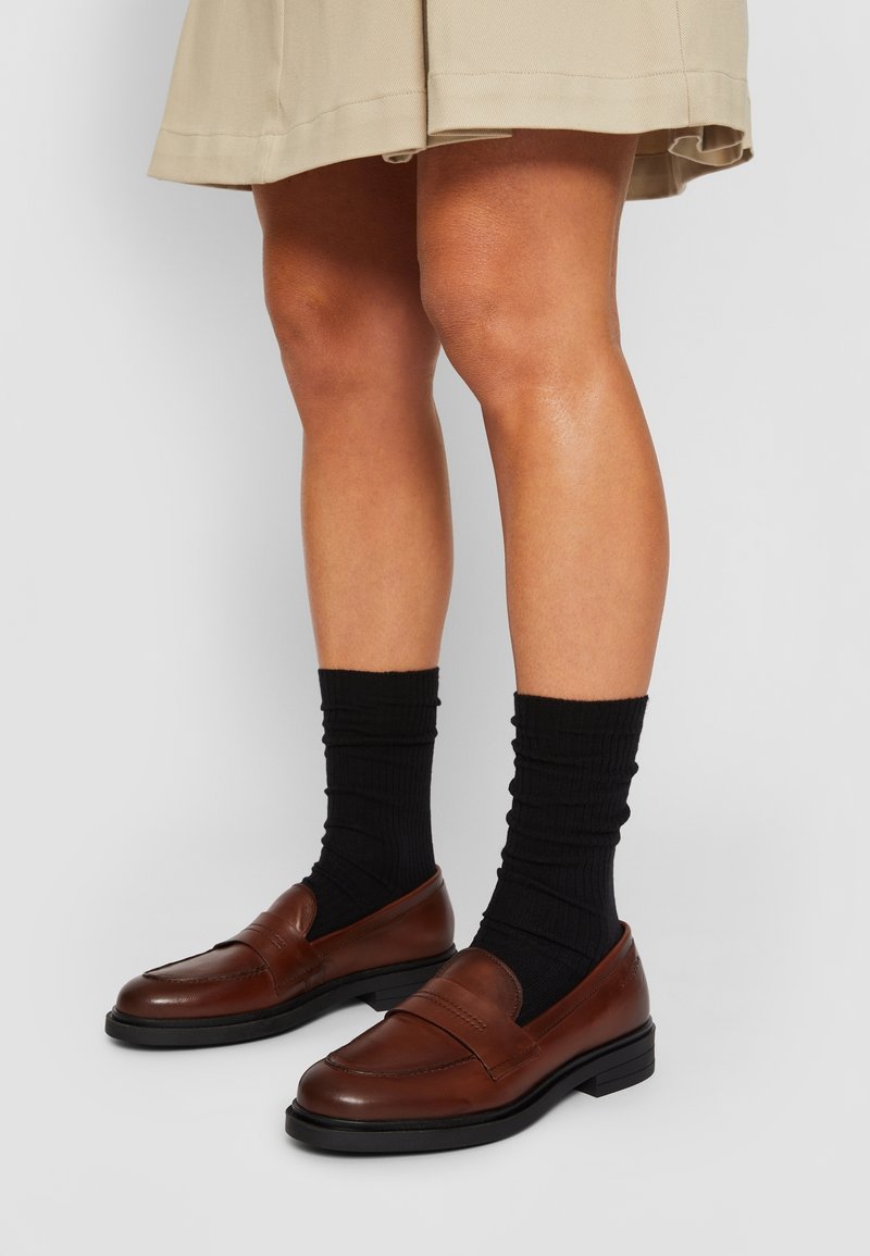Mocassins en cuir marron avec une finition lisse et des chaussettes noires côtelées extensibles, posés sur une jupe de couleur neutre. Gros plan sur les chaussures.