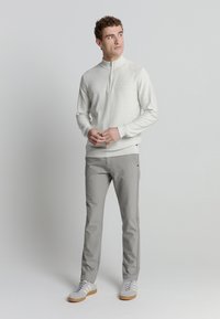 Lichtgrijze ritspullover, beige langslavige basislaag, grijze slim-fit broek en grijze sneakers met witte strepen. Eenvoudige, strakke lijnen.