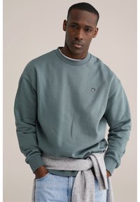 Zeleni bombažna jopa z okroglim izrezom in rebrastimi manšetami. Siva ruta zavezana okoli pasu, v kombinaciji z modrimi kavbojkami.