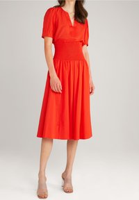 Robe en coton rouge avec des manches bouffantes courtes et un décolleté en V. Elle présente une taille smockée et une jupe évasée, accompagnée de sandales à talons transparents.