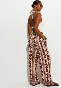 Mujer con top halter blanco, pantalones anchos color crema con patrones florales rojos, sandalias negras, gafas de sol, pendientes de aro dorados y pulseras.
