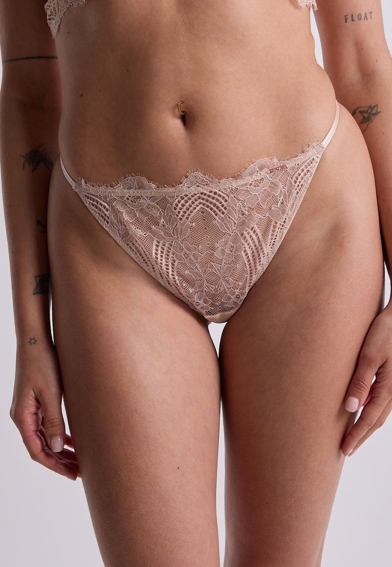 Culotte en dentelle couleur nude avec des bords festonnés, un motif floral et une couverture minimale. Fabriquée en matériau doux et transparent.