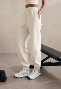 Kremfargede joggebukser med elastiske mansjetter og sidelommer, paret med lysegrå joggesko med blå detaljer og netting.