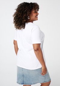 Witte katoenen T-shirt met korte mouwen en geborduurd hartdetail op de schouders, gecombineerd met een lichte spijkerrok. Krullend haar zichtbaar.