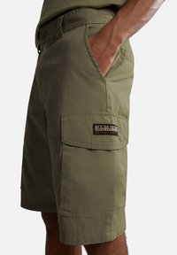 Pantaloni cargo verde oliva con tasche laterali, dotati di chiusura frontale con bottone, passanti per cintura e una toppa con logo sulla coscia. Tessuto leggero.