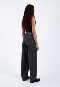 Breda svarta denimjeans med hög midja, med bakfickor och ett märkesmärke på midjan. Modellen har på sig en åtsittande svart topp och svarta skor.