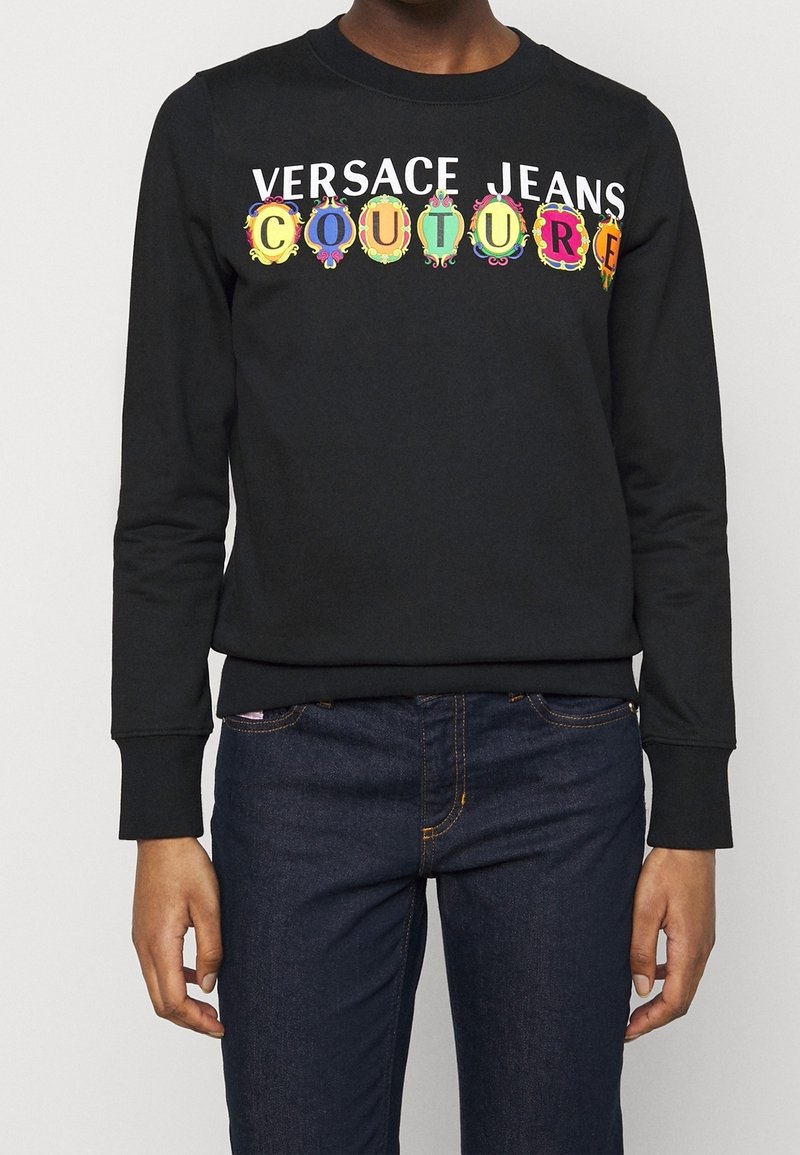 Personne portant un sweat-shirt noir avec le texte "Versace Jeans Couture", des lettres colorées encadrant "COUTURE", associé à un jean bleu foncé.