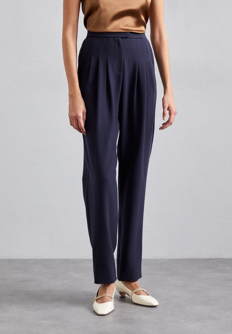 Navy high-waisted trousers with pleats, tailored fit, smooth fabric, paired with cream-colored low-heeled shoes featuring a gold accent.