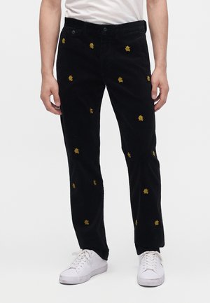 Pantaloni di velluto a coste neri con piccoli loghi gialli ricamati, indossati con scarpe da ginnastica bianche e una camicia bianca.