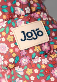Tessuto imbottito con un motivo floreale in tonalità di rosa, giallo e verde. Presenta un'etichetta color crema con "JoJo" in blu scuro.