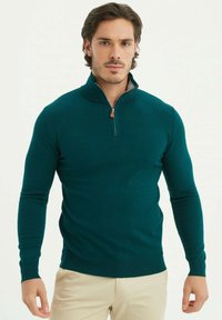Pull en maille teal avec un col haut, une fermeture éclair sur le devant et des poignets côtelés. Fabriqué à partir d'un tissu doux, il présente une coupe ajustée et une texture lisse.