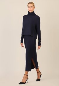 Navy-Blau gestrickter Pullover mit hohem Kragen, kombiniert mit einem passenden gerippten Rock mit seitlichen Schlitzen, ergänzt durch schwarze Stiletto-Absätze.