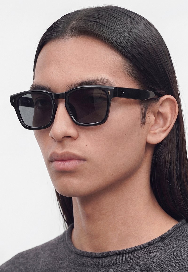 Personne aux longs cheveux noirs portant des lunettes de soleil rectangulaires noires et un haut gris à col rond sur un fond uni.