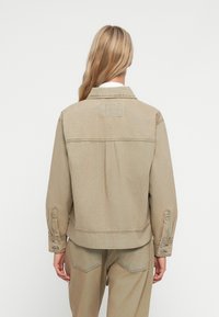 Beige denimjakke med avslappet passform, ryggsøm og to-knapps justerbare mansjetter. Har en rektangulær lapp på øvre del av ryggen.