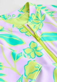 Tuvo plāns ar lavandas audumu, uz kura ir spilgti zaļi un tirkīza ziedu raksti, un daļēji aizvērts zaļš rāvējslēdzējs.