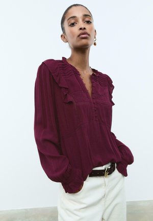 Blouse froncée bordeaux avec des détails texturés, fermeture à boutons et manches longues. Associée à un pantalon clair et une ceinture noire.