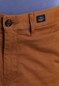 Pantaloni di cotone marrone con una texture liscia, dotati di un bottone nero, passanti per la cintura e un'etichetta che mostra i dettagli del marchio e del paese d'origine.