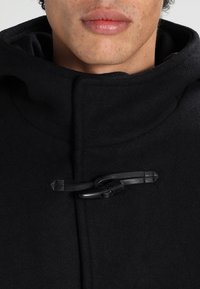 Abrigo de lana negro con un cuello alto, cierre frontal de botones con cordón negro y herrajes, y una textura suave.