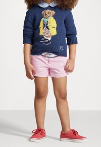 Marineblauer Strickpullover mit einem Bärenmotiv, der einen gelben Regenmantel trägt, kombiniert mit rosa Shorts und roten Turnschuhen mit weißen Sohlen.