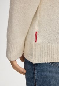 Primo piano di una mano vicino all'orlo di un maglione in knit color crema con un'etichetta rossa "JOOP! JEANS", abbinato a un paio di jeans in denim blu.