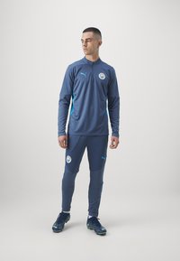 Tuta sportiva blu scuro con accenti blu chiaro, caratterizzata da un top con zip a un quarto e pantaloni aderenti, completata da scarpe sportive abbinate.