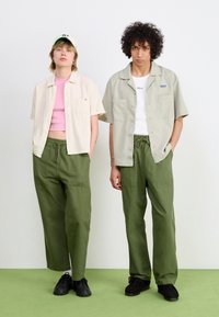 Deux individus portent des tenues décontractées : l'un est habillé d'une chemise à rayures beige à manches courtes, d'un haut rose et d'un pantalon vert ; l'autre porte une chemise grise, un t-shirt blanc et un pantalon vert.