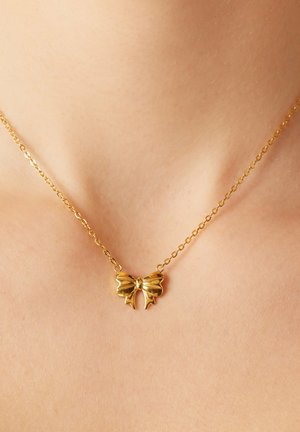 Gouden ketting met een vlinder-vormige hanger met gestructureerde vleugels en een centraal strikelement, liggend op een blote nek.