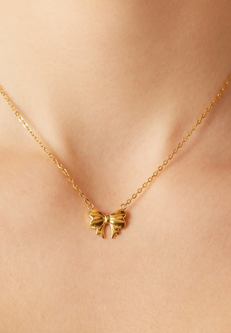 Gouden ketting met een vlinder-vormige hanger met gestructureerde vleugels en een centraal strikelement, liggend op een blote nek.
