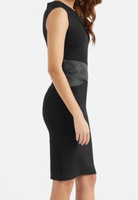 Svart tajt klänning med korta ärmar, med en midja i läder och slät textur. Knäl ång klippning, minimalistisk design.