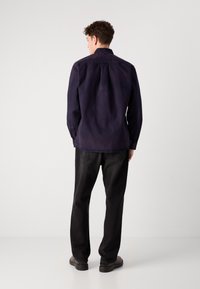 Cămașă din bumbac bleumarin cu manșete cu nasturi, detaliu la spate, tăietură dreaptă, purtată cu pantalonii din denim negri și pantofi închiși la culoare.