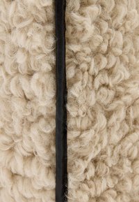 Beige, gestructureerde stof met krullende vezels, omrahmd door een dunne zwarte leren rand, die een zachte en pluizige uitstraling geeft.