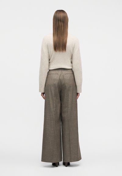 Γυναίκα με μακριά ίσια μαλλιά που φοράει μπεζ πουλόβερ και καφέ παντελόνι ψηλόμεσο, φαρδύ, που στέκεται με την πλάτη προς τα εμπρός σε λευκό φόντο.