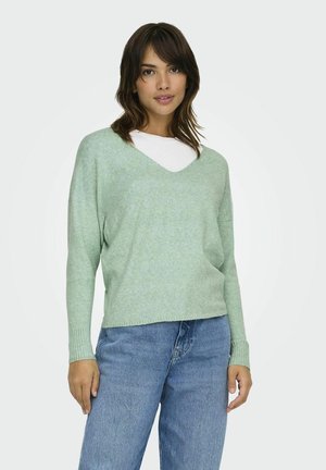 Pull en tricot vert clair avec un col en V, coupe décontractée et poignets côtelés, superposé à une chemise blanche, associé à un jean bleu.