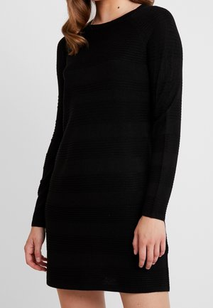 Frau trägt ein eng anliegendes langärmliges schwarzes Kleid mit subtiler horizontaler Rippstruktur, steht vor einem schlichten hellen Hintergrund.