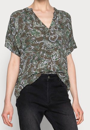 Woman wearing a short-sleeve olive green paisley blouse tucked into high-waisted black jeans, shown from shoulders to hips.