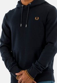 Sudadera navy con un bolsillo tipo canguro en la parte frontal, cuenta con cierre de cordón, logo dorado en el pecho y puños acanalados naranjas en contraste.